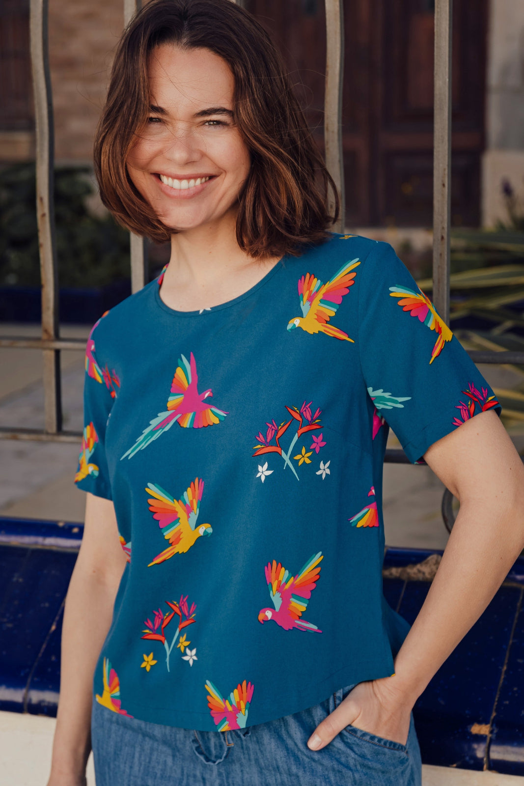 A woman smiling and wearing a teal t-shirt with a pattern of colorful rainbow parrots.