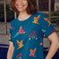 A woman smiling and wearing a teal t-shirt with a pattern of colorful rainbow parrots.