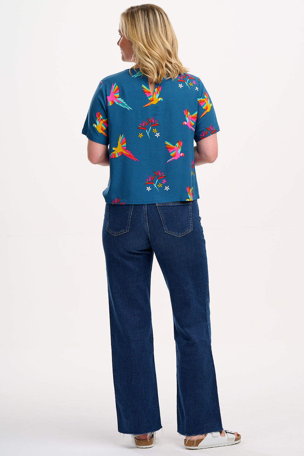 A woman wearing a teal t-shirt with a pattern of rainbow parrots, paired with jeans.
