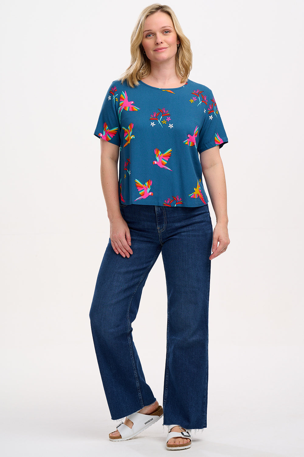A woman wearing a teal t-shirt with a pattern of rainbow parrots, paired with jeans.