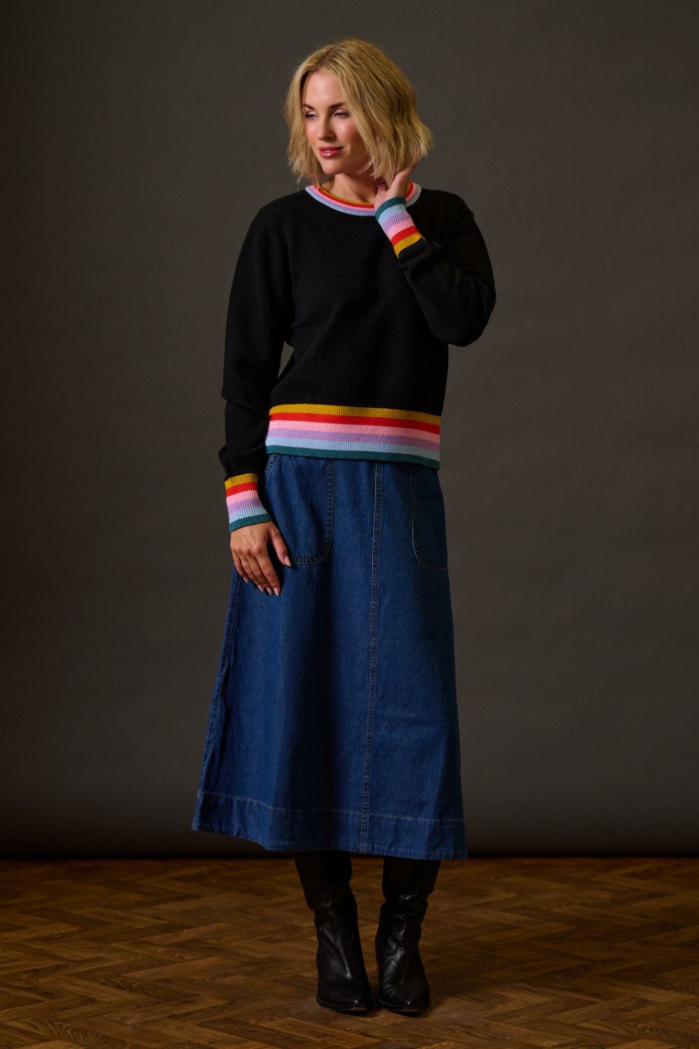 Woman wearing a black sweater with colorful stripes and a blue denim skirt against a plain background