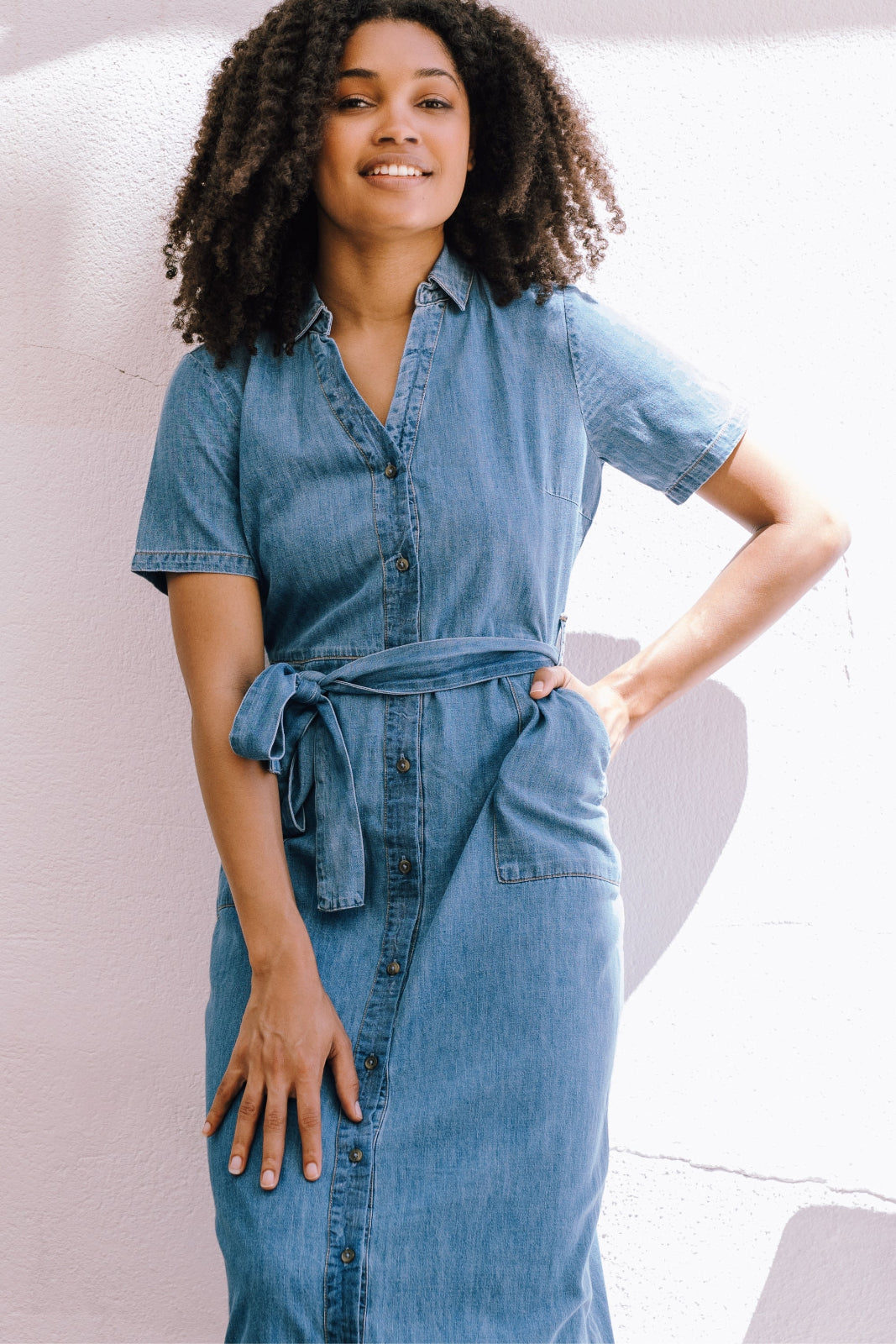 A woman wearing a mid blue denim midi shirt dress with a collar, short sleeves, waist belt tie, and patch pockets.