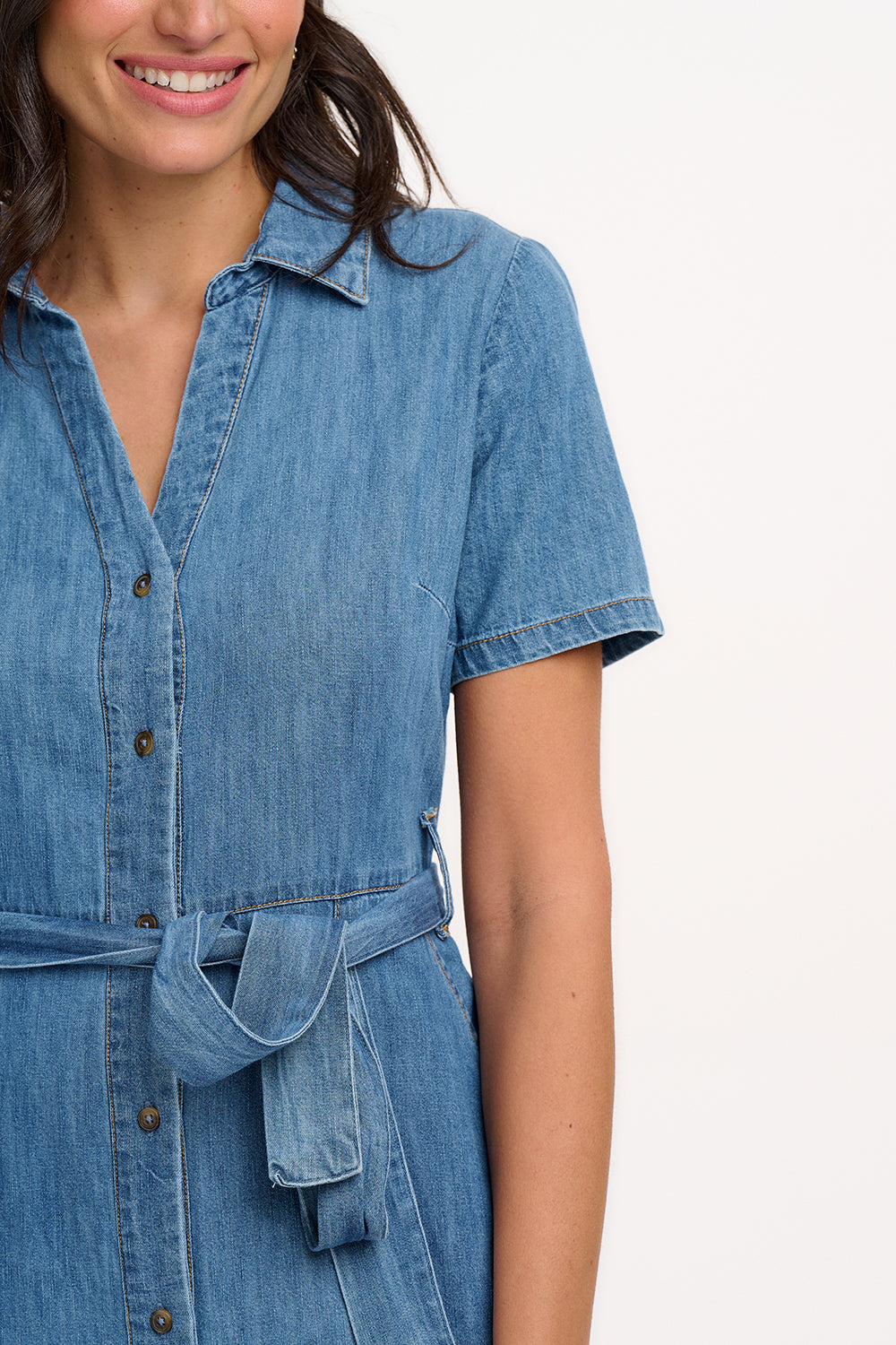A woman standing in front of a white background wearing a mid blue denim midi shirt dress with short sleeves, a collared neckline, and a waist belt tie. She has patch pockets on the front of the dress.