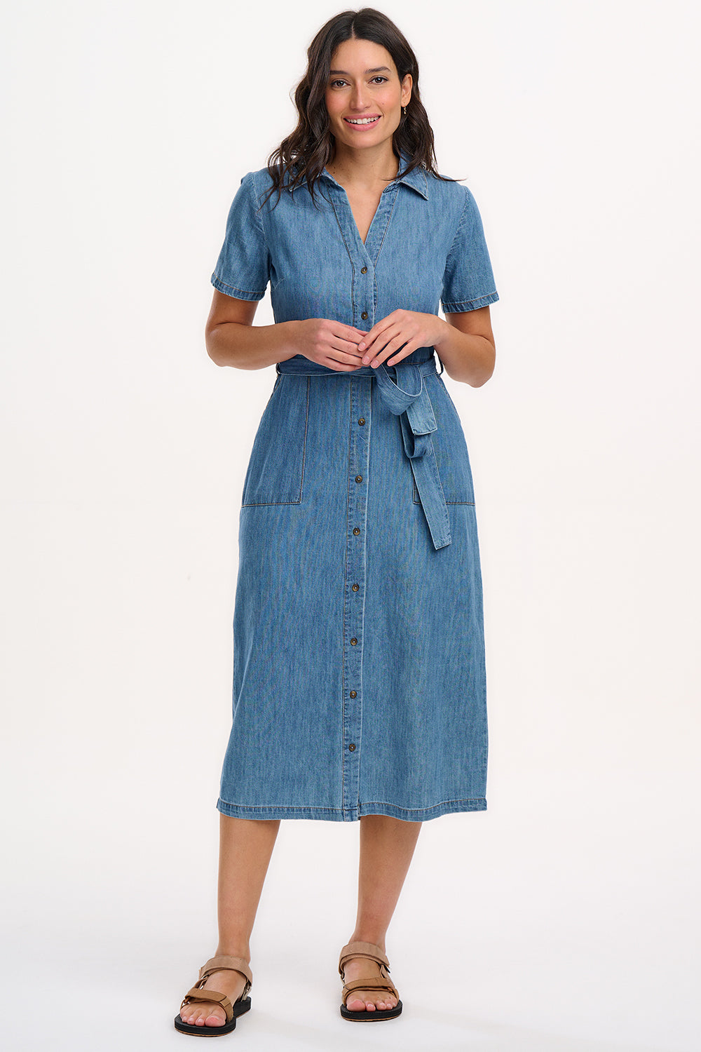 A woman standing in front of a white background wearing a mid blue denim midi shirt dress with short sleeves, a collared neckline, and a waist belt tie. She has patch pockets on the front of the dress.