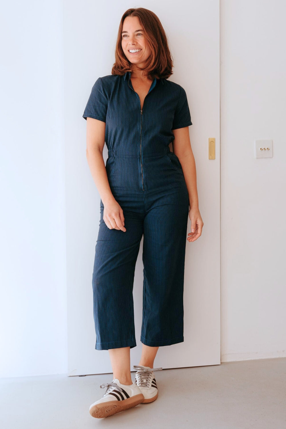 Woman wearing a dark blue jumpsuit standing against a white wall.
