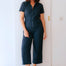 Woman wearing a dark blue jumpsuit standing against a white wall.