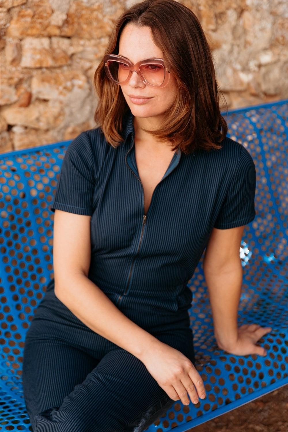 Woman wearing sunglasses sitting on a blue bench against a stone wall.