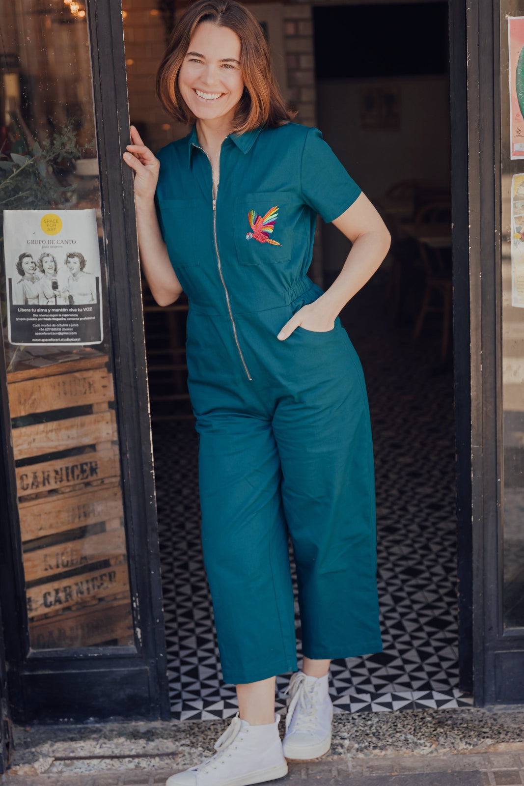 A woman standing in a doorway wearing a teal short sleeve jumpsuit with a rainbow parrot embroidered on the chest area, paired with white sneakers.
