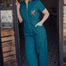 A woman standing in a doorway wearing a teal short sleeve jumpsuit with a rainbow parrot embroidered on the chest area, paired with white sneakers.