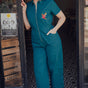 A woman standing in a doorway wearing a teal short sleeve jumpsuit with a rainbow parrot embroidered on the chest area, paired with white sneakers.