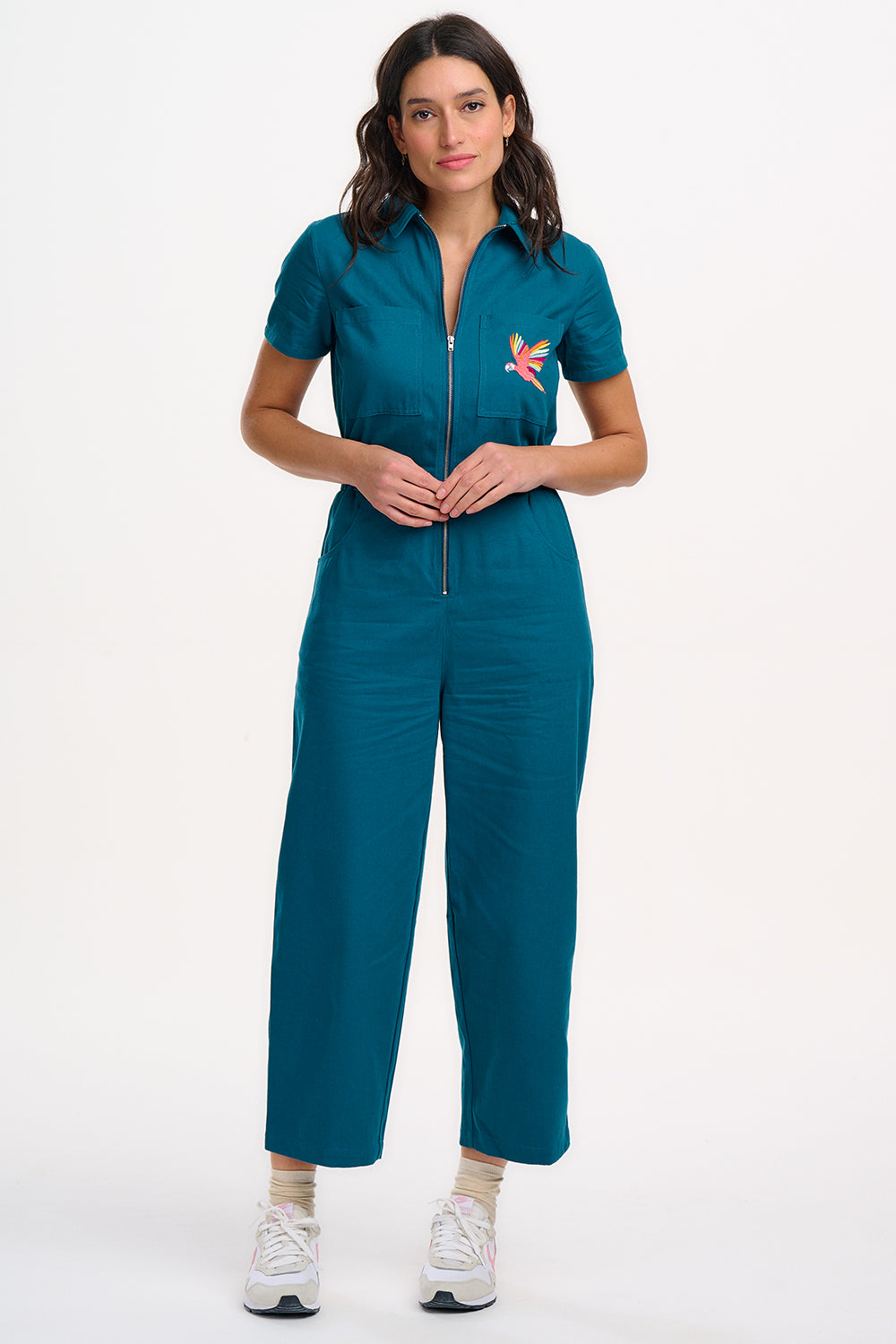 A woman modeling a teal-colored jumpsuit with short sleeves, a front zip, and wide flared legs. The jumpsuit features a pocket on the chest area and is embroidered with a rainbow parrot design.