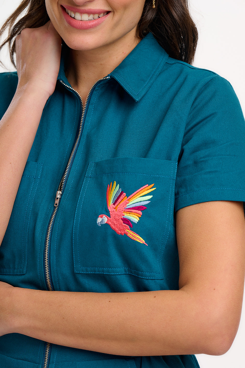 A woman modeling a teal-colored jumpsuit with short sleeves, a front zip, and wide flared legs. The jumpsuit features a pocket on the chest area and is embroidered with a rainbow parrot design.