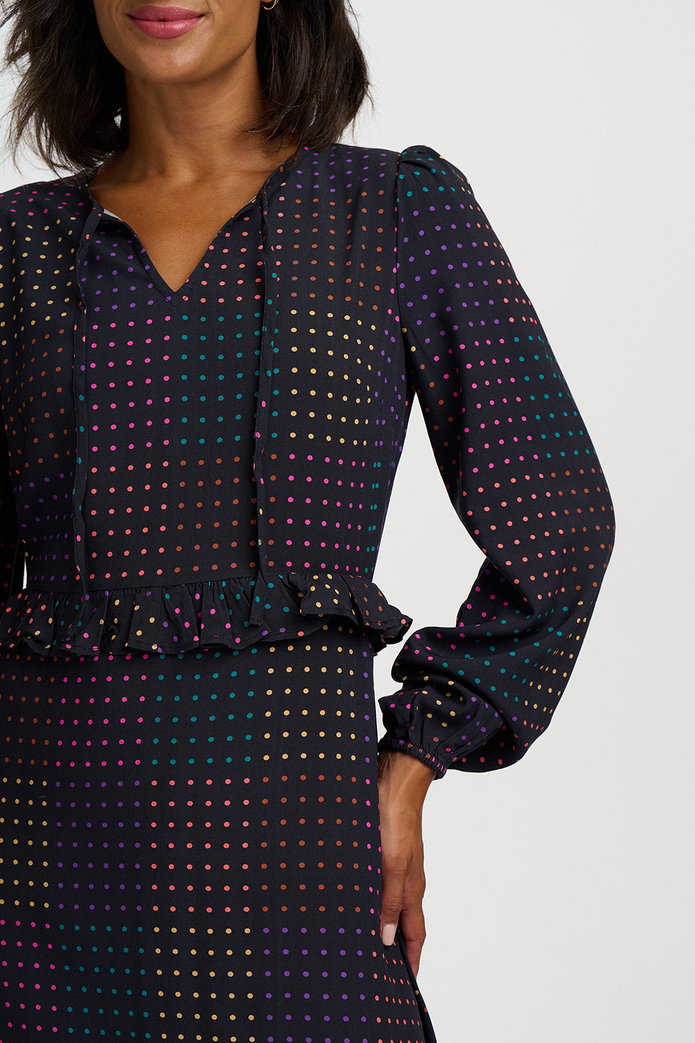 A close-up of a model wearing a long-sleeve V-neck dress with a dark base and a dense multicolor polka dot pattern, featuring a ruffled waistband.
