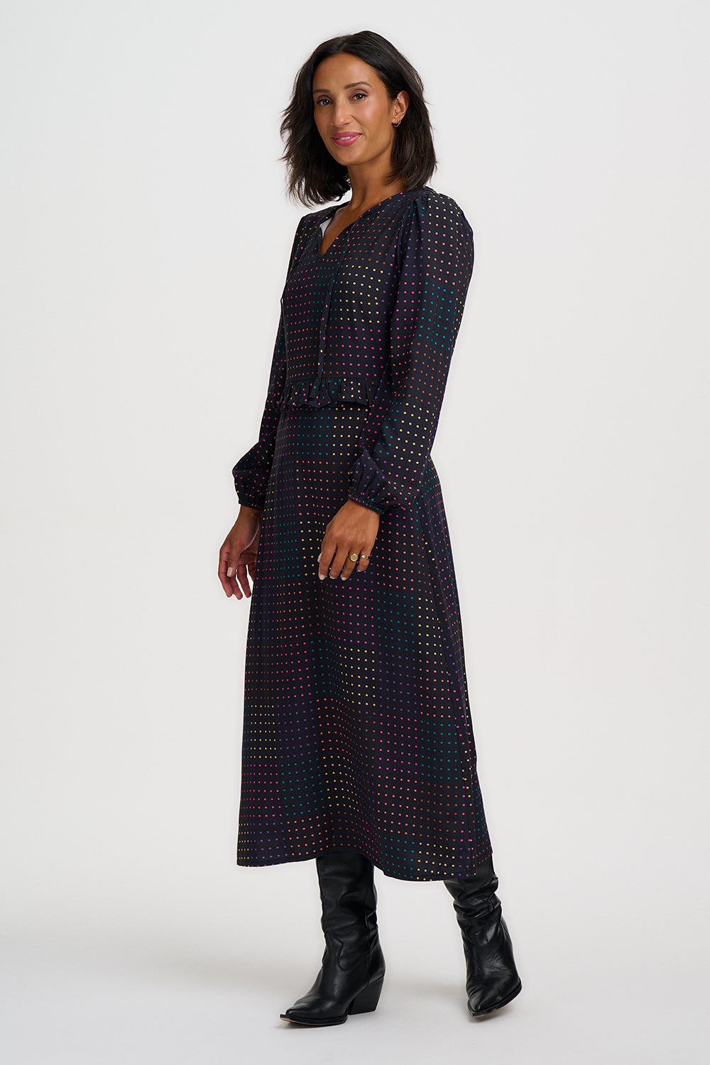 A woman modeling a long-sleeved midi dress with a dark navy and black base and a small, dense multicolor geometric pattern. She is wearing black leather ankle boots against a plain light gray studio background.
