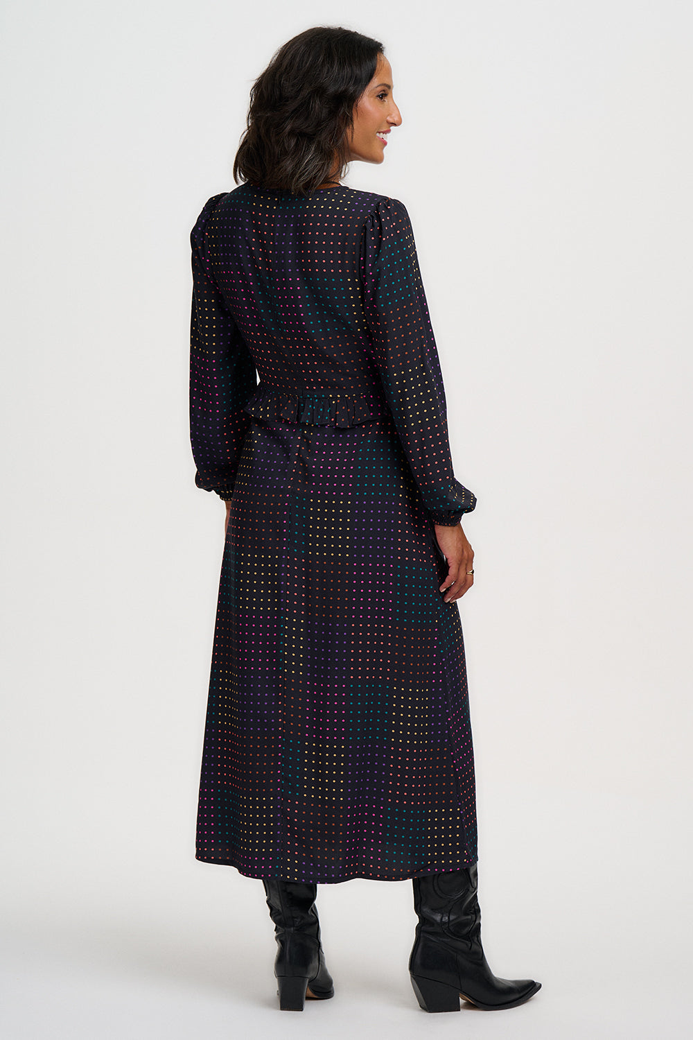 A woman shown from the back wearing a long-sleeved midi dress with a dark base and a small repeating geometric pattern in light contrasting dots. She is wearing black ankle boots against a plain light gray studio background.