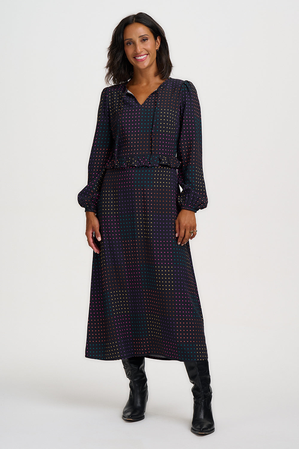 A woman modeling a long-sleeve midi dress with a dark navy and black base and a dense small gold geometric pattern, featuring a matching waist belt, against a plain light gray studio background.