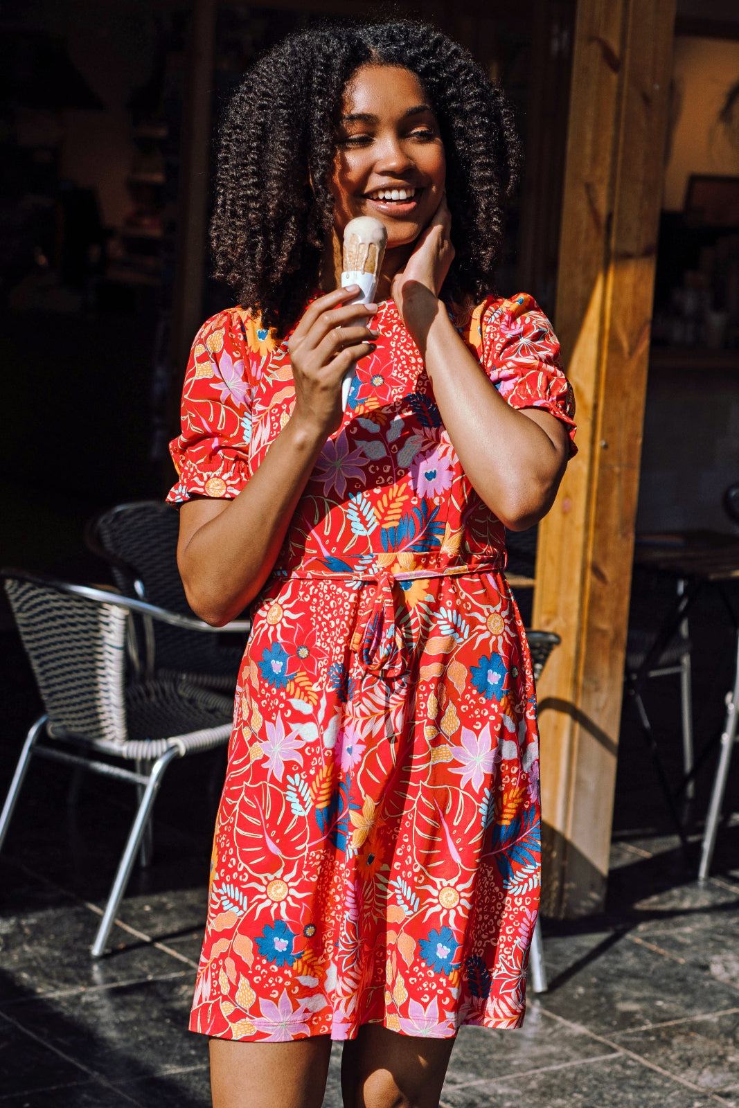 A woman wearing a red fit and flare jersey mini dress with a round neck, skinny belt, and short shirred sleeves featuring a colorful tropical leaf and flower print.