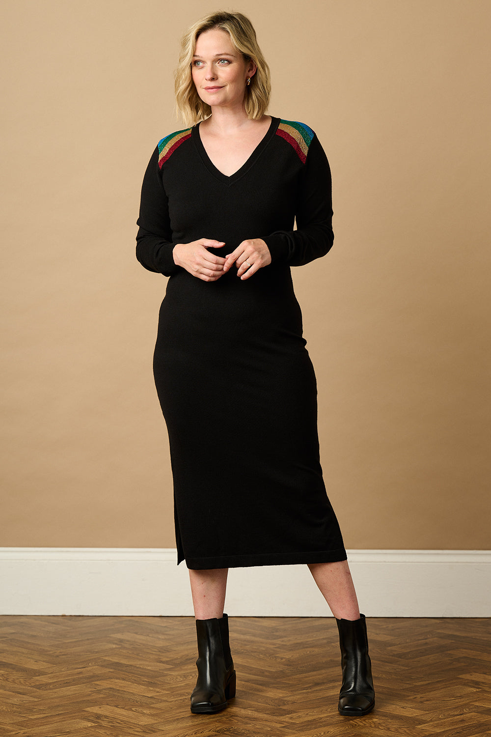 A woman standing wearing a long-sleeved, V-neck, black knit midi dress with a split side hem, paired with black boots.
