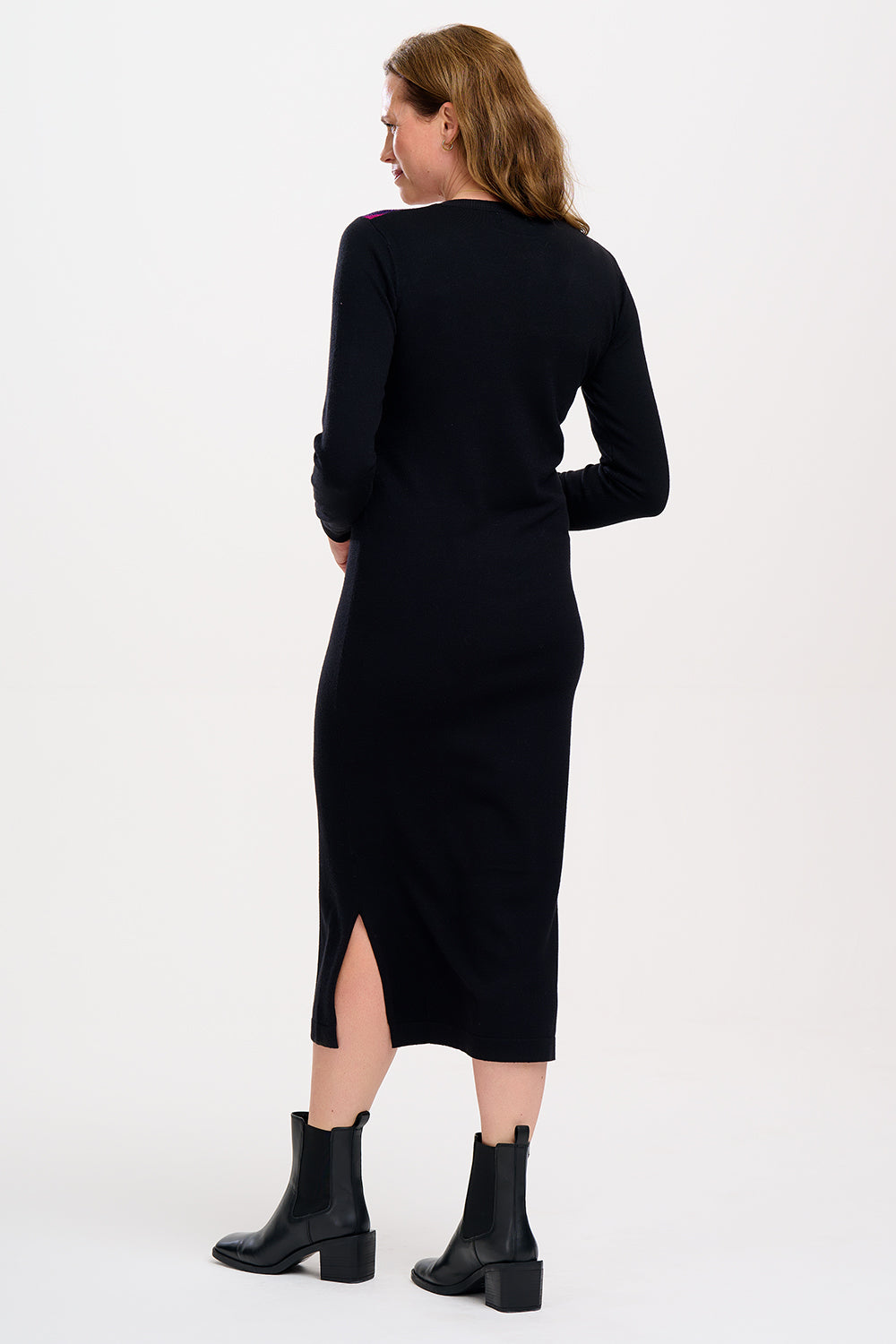 A woman standing wearing a long-sleeved, V-neck, black knit midi dress with a split side hem, paired with black boots.