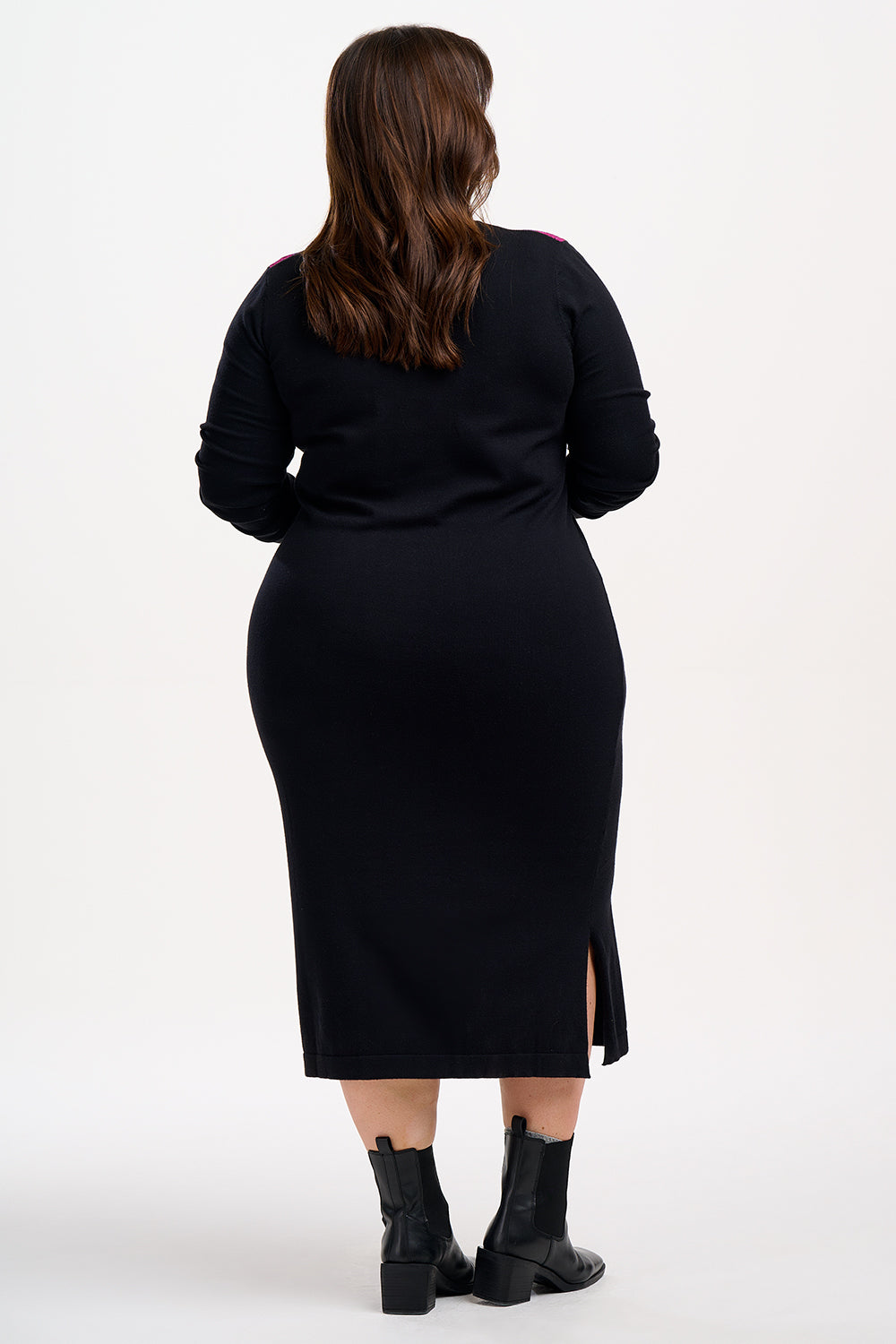 A woman standing wearing a long-sleeved, V-neck, black knit midi dress with a split side hem, paired with black boots.