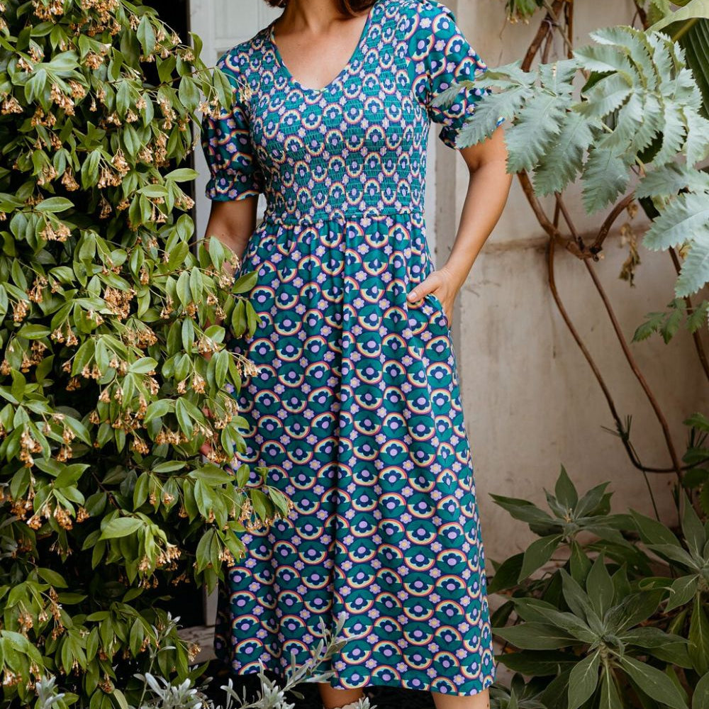 Woman wearing a blue patterned dress standing outdoors with greenery around.