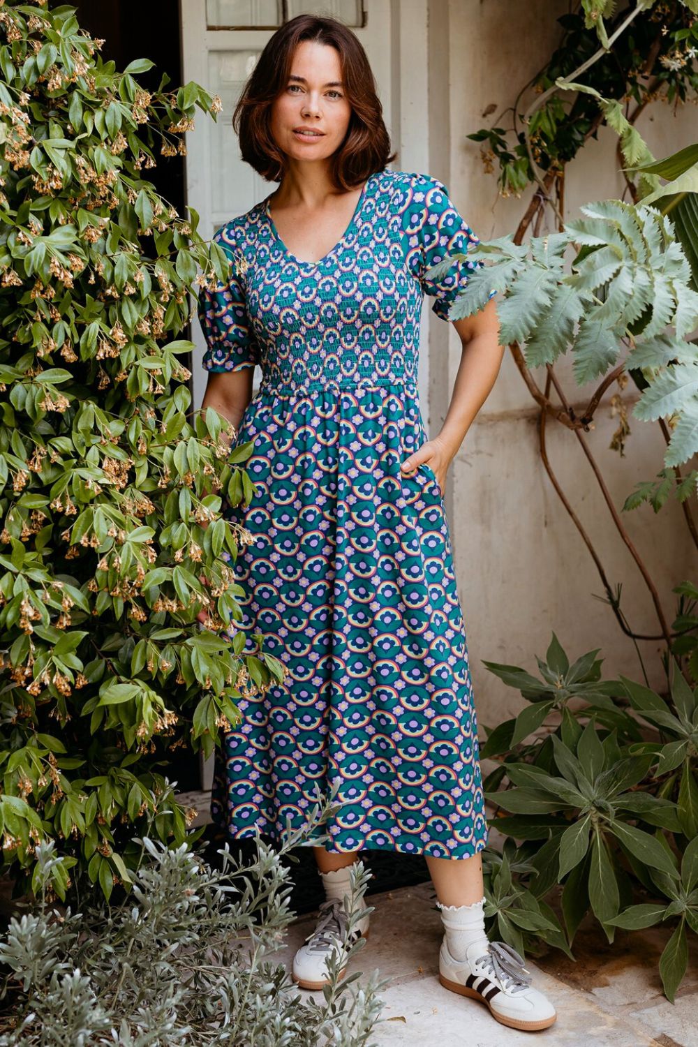 Woman wearing a blue patterned dress standing outdoors with greenery around.