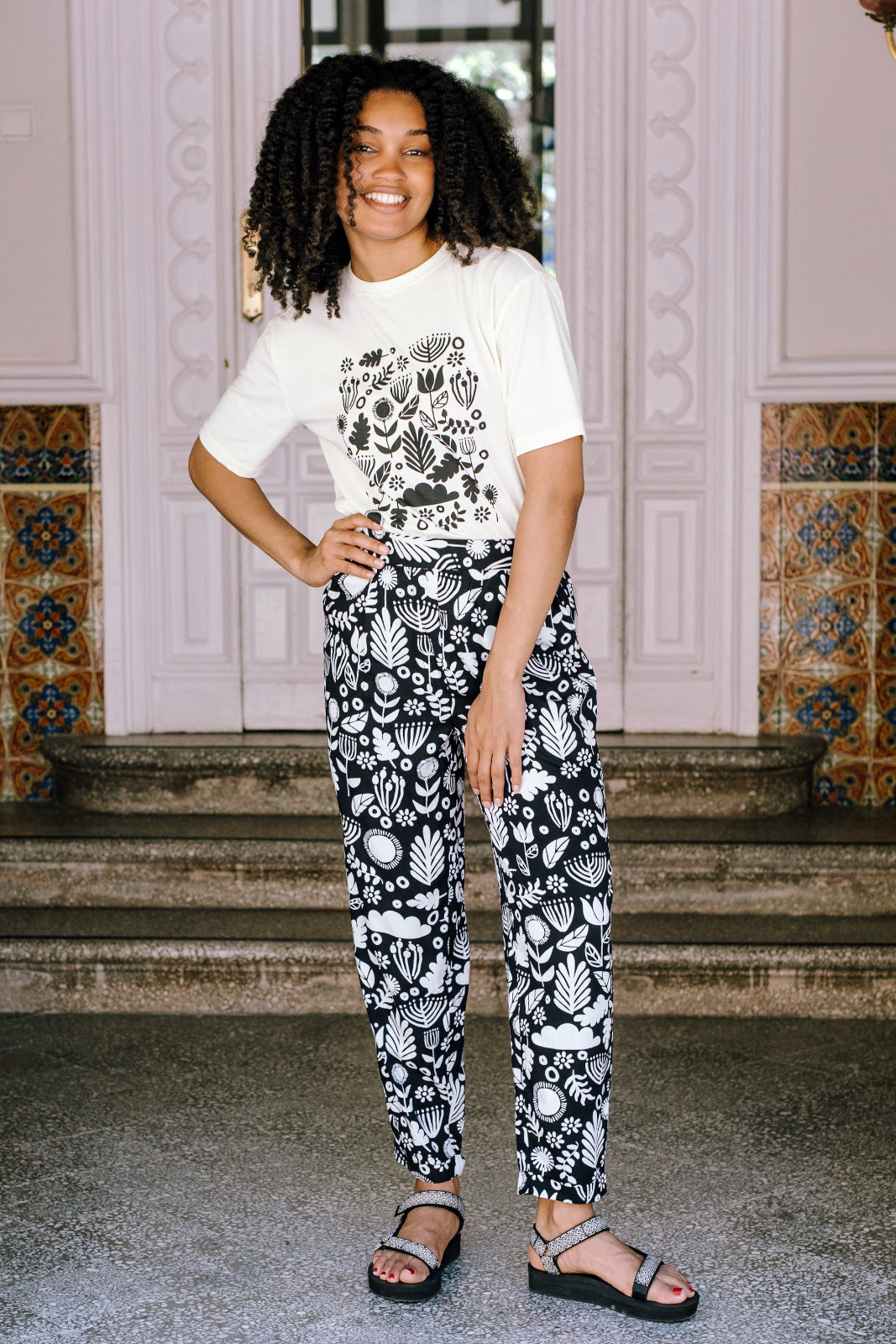 A person standing and smiling, wearing a white top with black floral folk print trousers, with turned-up hem and elasticated waist, paired with black strappy sandals.