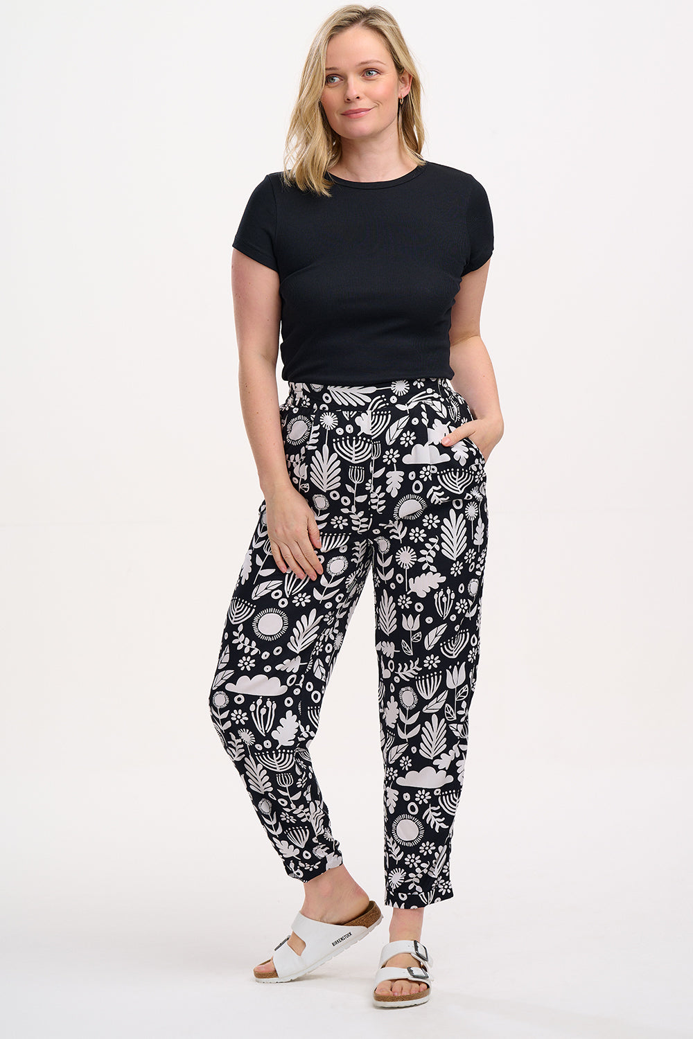 A woman standing, wearing a black top and Debbs trousers with a monochrome floral pattern.