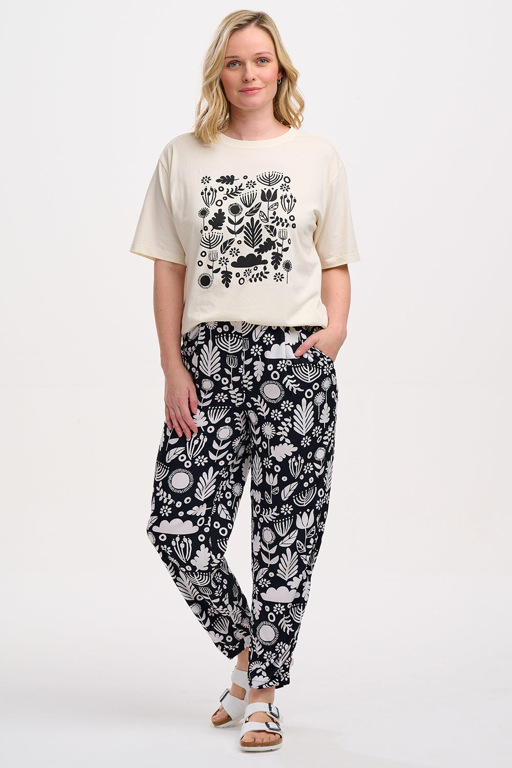 A woman standing, wearing a black top and Debbs trousers with a monochrome floral pattern.