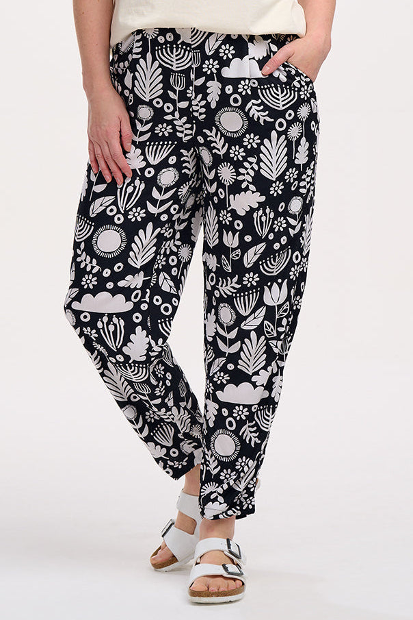 A woman standing, wearing a black top and Debbs trousers with a monochrome floral pattern.