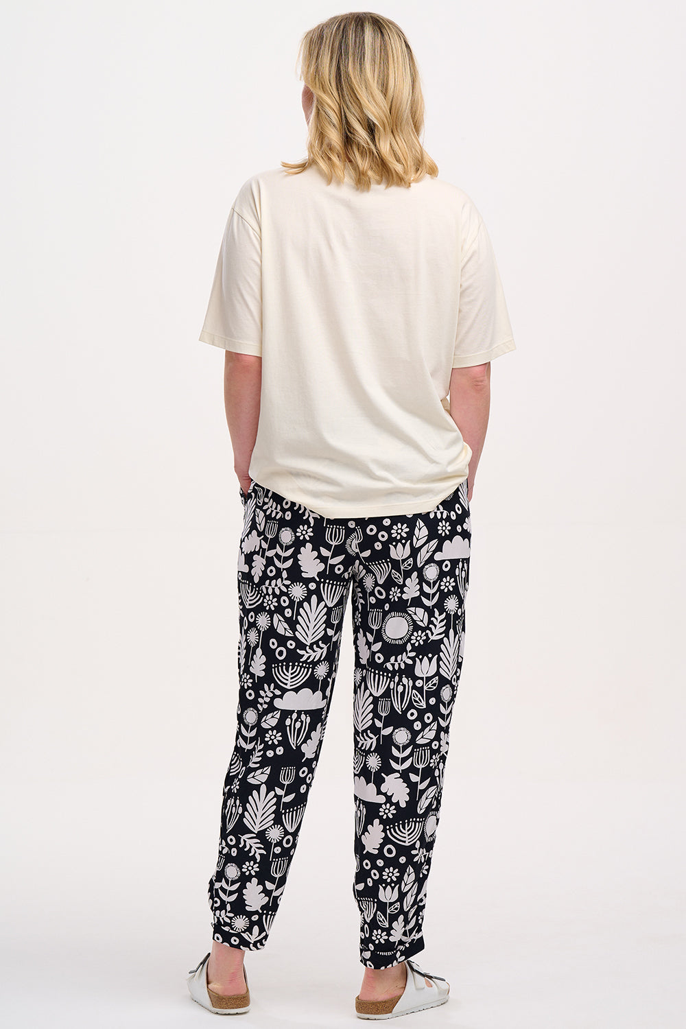 A woman standing, wearing a black top and Debbs trousers with a monochrome floral pattern.
