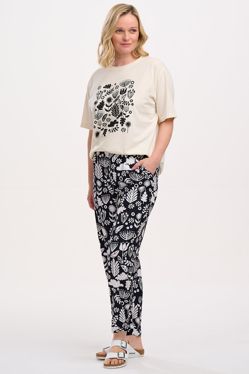 A woman standing, wearing a black top and Debbs trousers with a monochrome floral pattern.