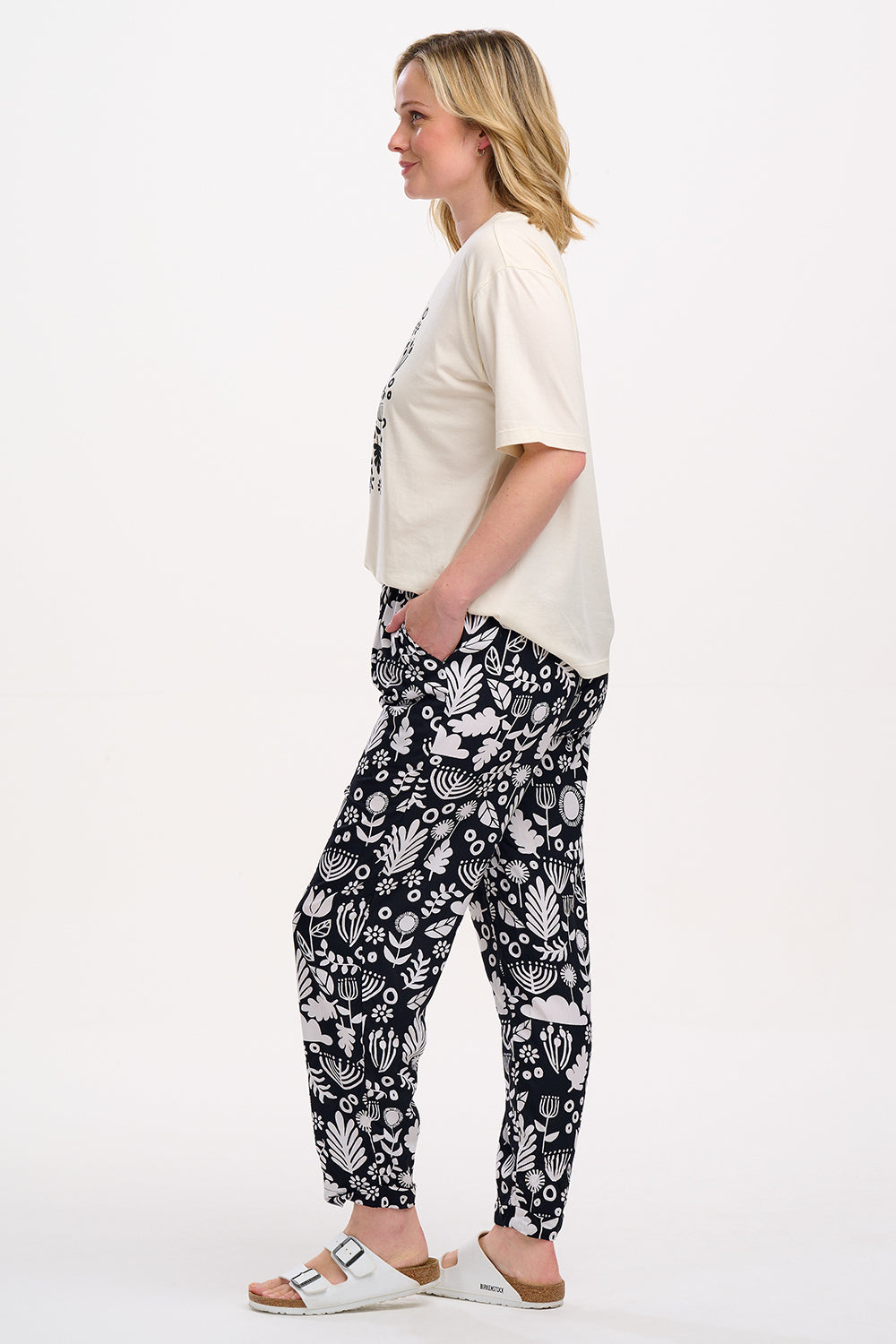 A woman standing, wearing a black top and Debbs trousers with a monochrome floral pattern.