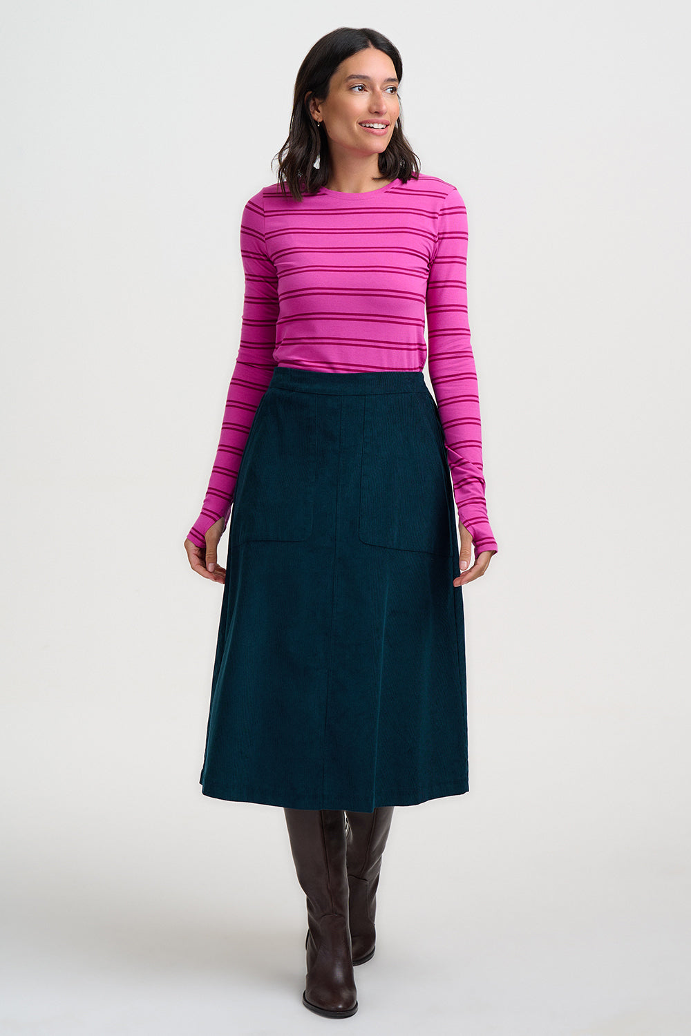 A woman modeling a bright pink and magenta striped long-sleeve top paired with a dark teal skirt and brown knee-high boots, standing against a plain light gray studio background.