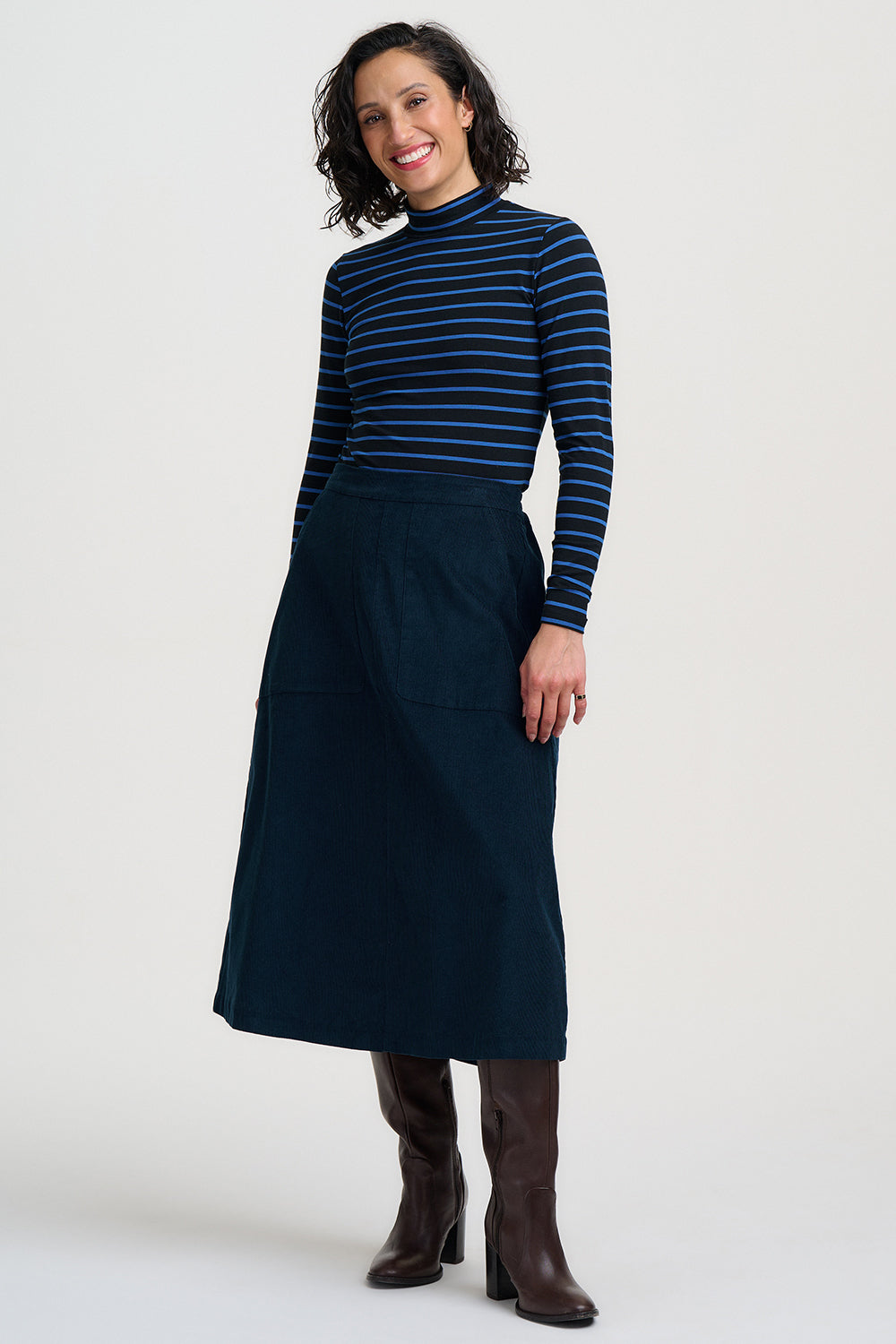 A woman modeling a navy blue mock-neck long-sleeve top with thin dark stripes, paired with a matching navy midi skirt and dark brown knee-high boots, against a light gray studio background.