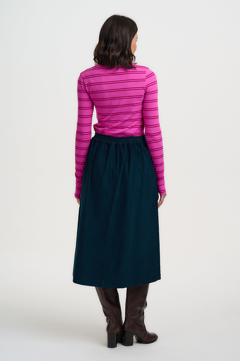 A woman viewed from the back wearing a bright pink and magenta striped long-sleeve top, a dark teal midi skirt, and brown knee-high boots, against a plain light gray studio background.