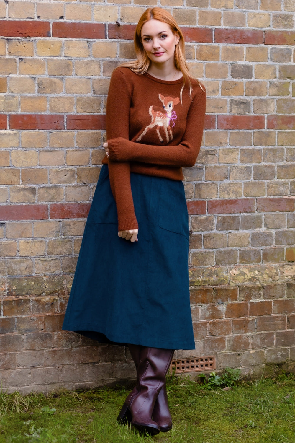 A woman stands against a brick wall wearing a rust-brown knit sweater with a white deer graphic, a dark teal midi skirt, and dark brown leather boots.