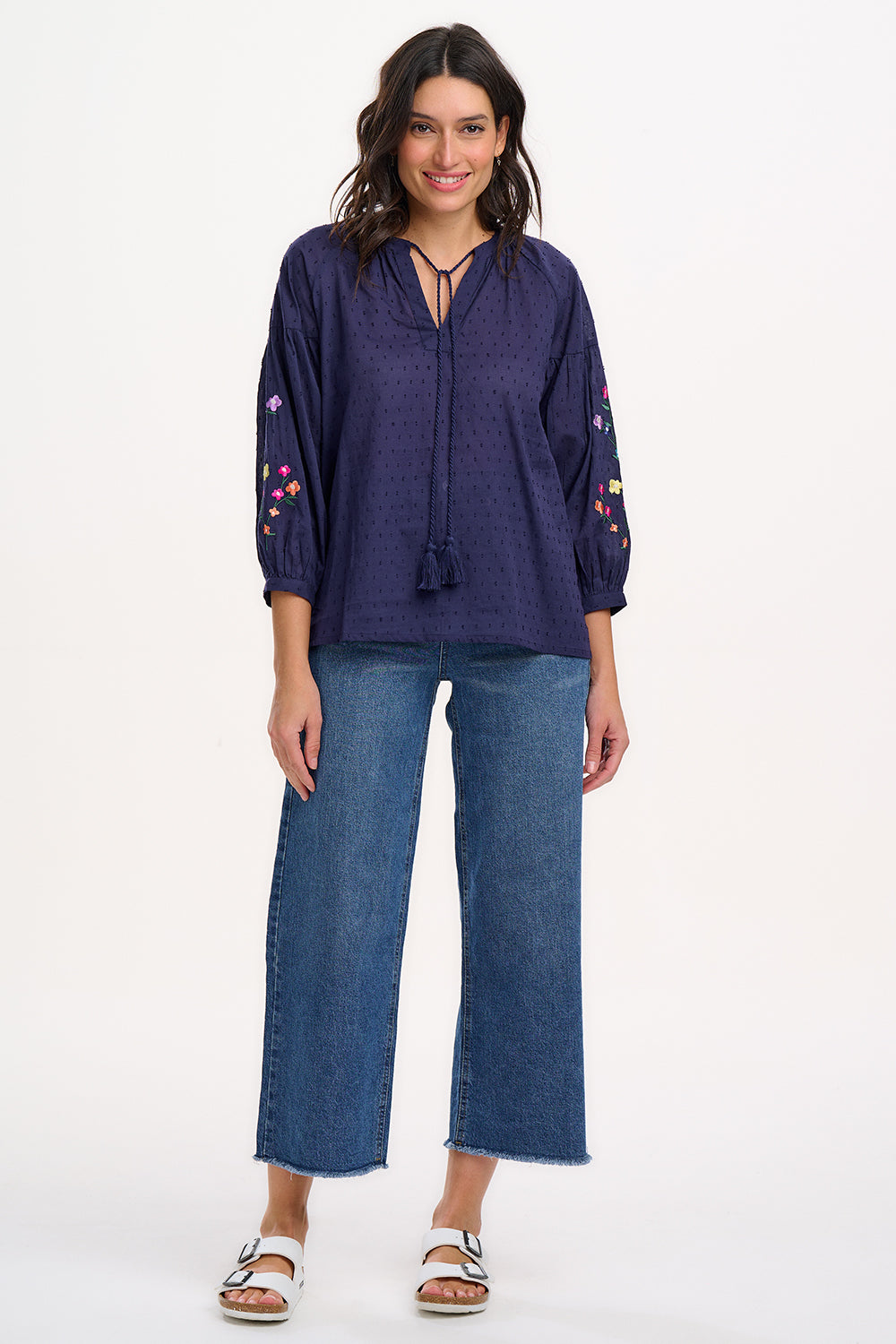 A woman wearing a navy blue boho top with embroidered flowers on the sleeves, featuring a deep V-neck with tassel ties and 3/4 length sleeves.