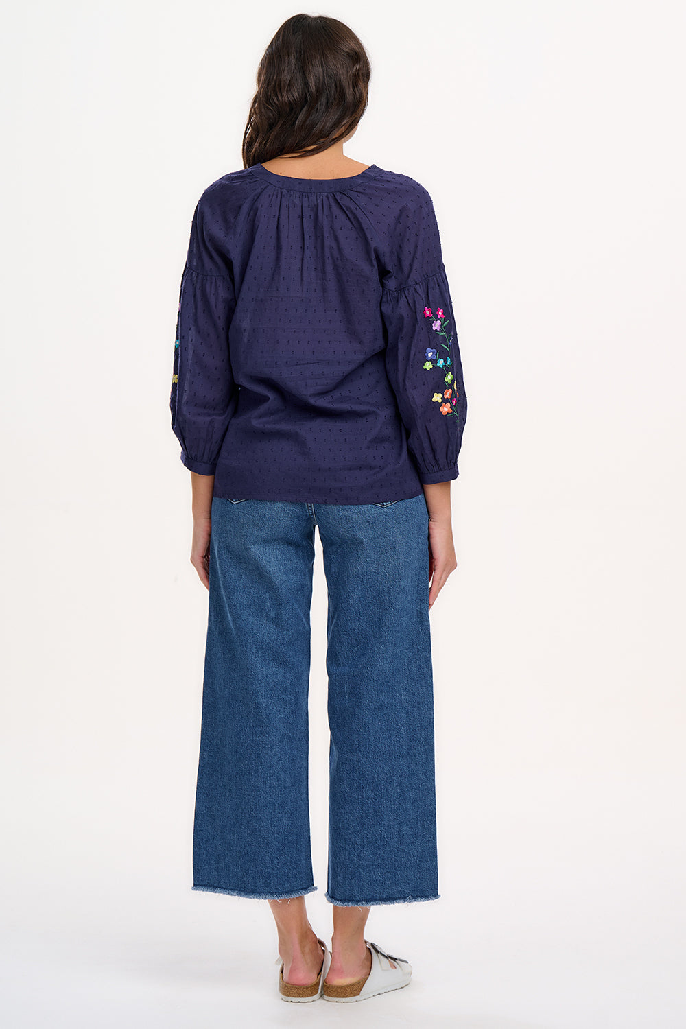 A woman wearing a navy blue boho top with embroidered flowers on the sleeves, featuring a deep V-neck with tassel ties and 3/4 length sleeves.