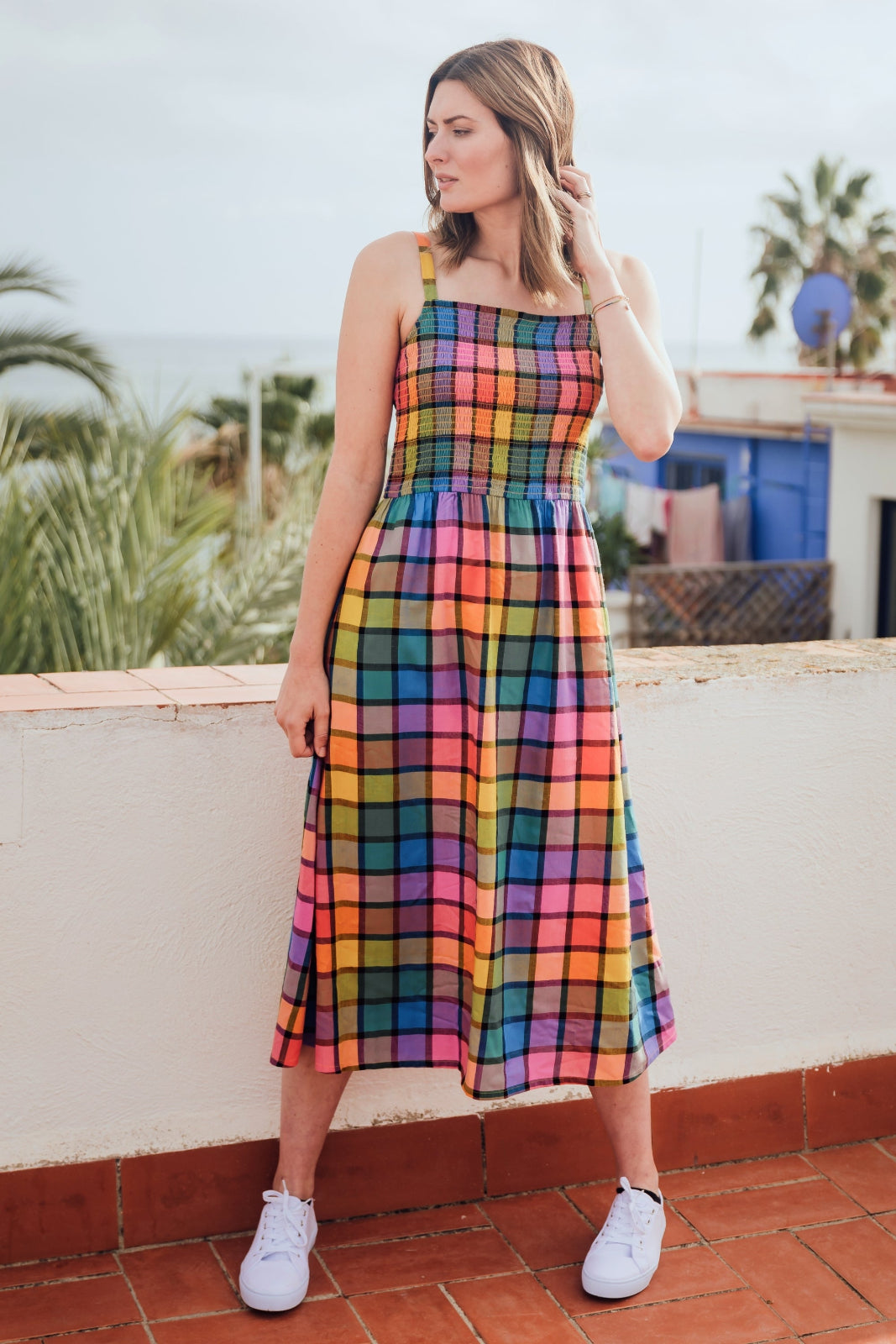 A woman wearing a multicolored checkered midi sundress with a shirred bodice and thin straps, in a rainbow check pattern.