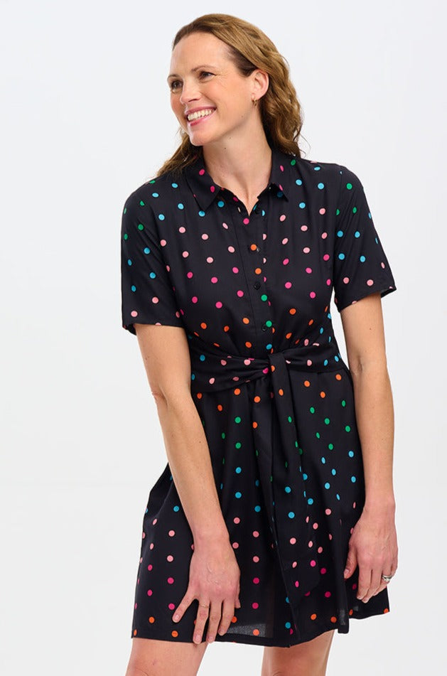 A woman wearing a knee-length black shirt dress with a multicolored polka dot pattern, short sleeves, and a tie-front belt.