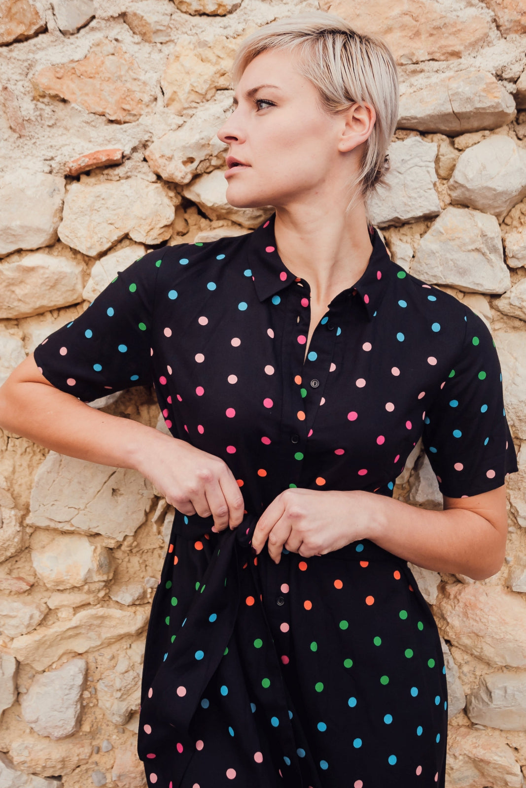 Woman wearing a black dress with colorful polka dots against a stone wall.
