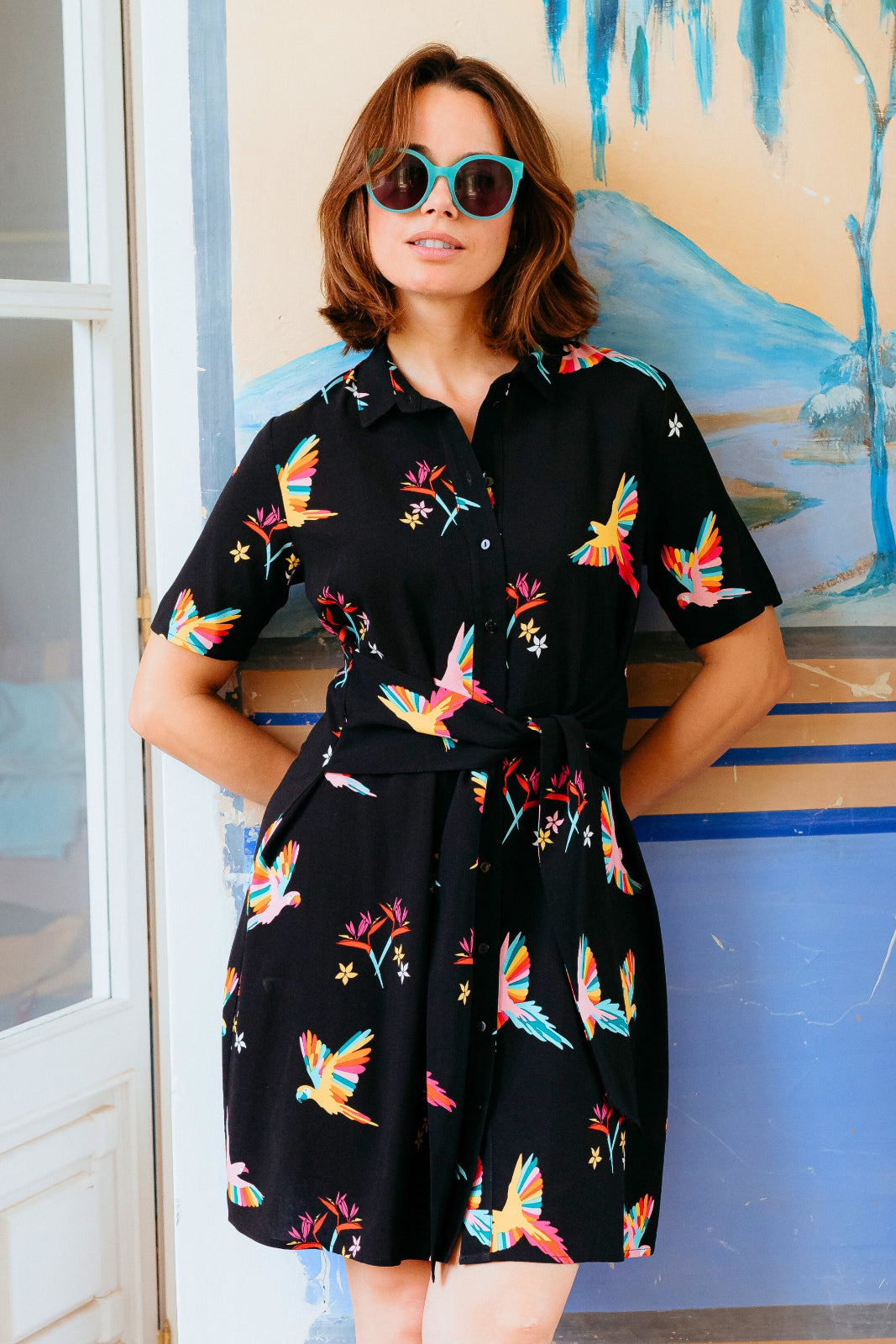 Woman wearing a black dress with colorful bird patterns in front of a colorful mural.