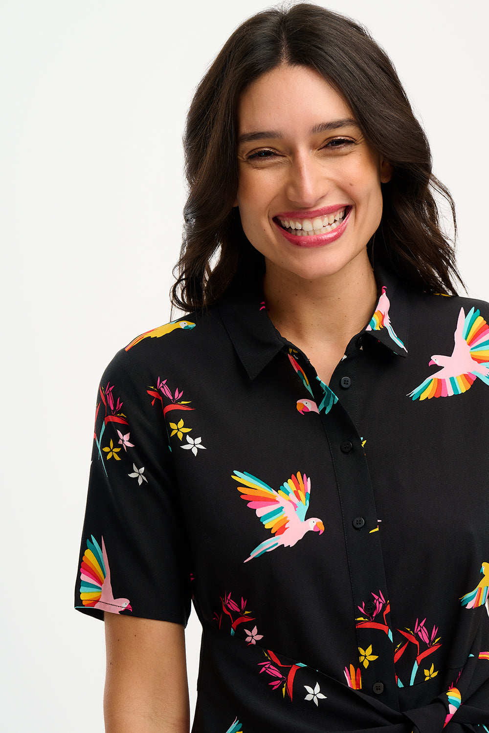 A model wearing a short-sleeved black shirt dress with a Tropical Parrots print, featuring a collar, button-up front, and a tie-front belt.