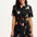 A model wearing a short-sleeved black shirt dress with a Tropical Parrots print, featuring a collar, button-up front, and a tie-front belt.