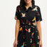 A model wearing a short-sleeved black shirt dress with a Tropical Parrots print, featuring a collar, button-up front, and a tie-front belt.