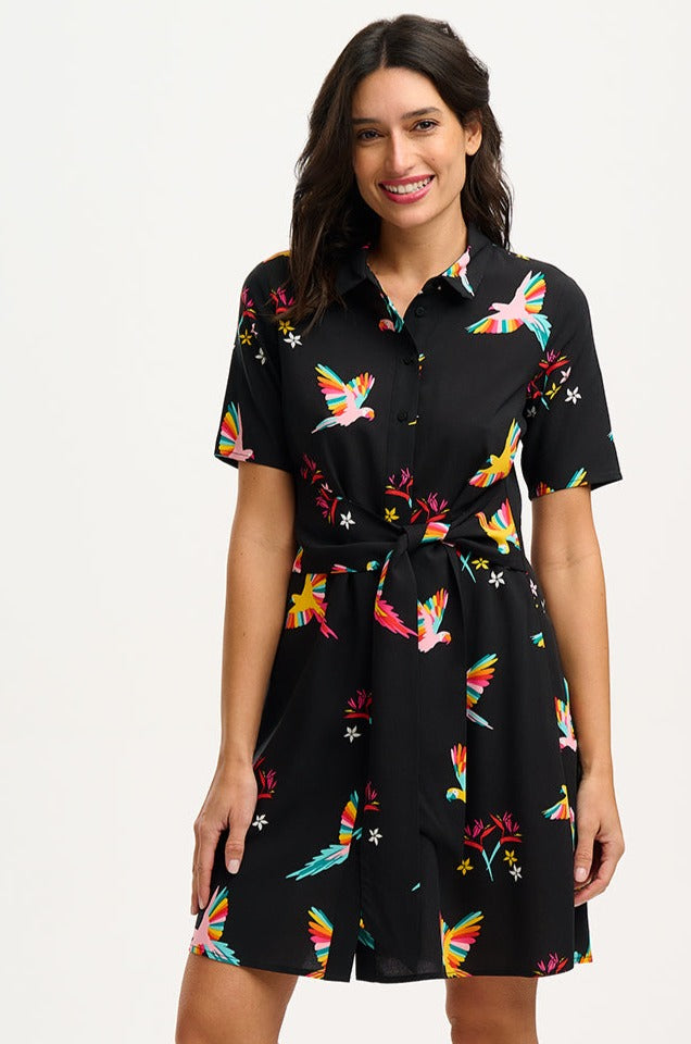 A model wearing a short-sleeved black shirt dress with a Tropical Parrots print, featuring a collar, button-up front, and a tie-front belt.