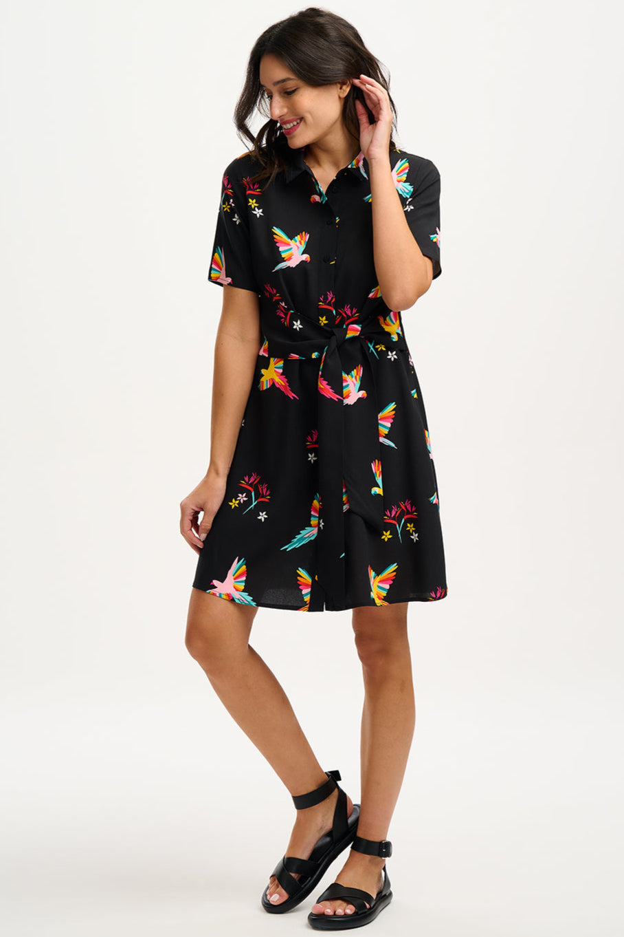 A model wearing a short-sleeved black shirt dress with a Tropical Parrots print, featuring a collar, button-up front, and a tie-front belt.