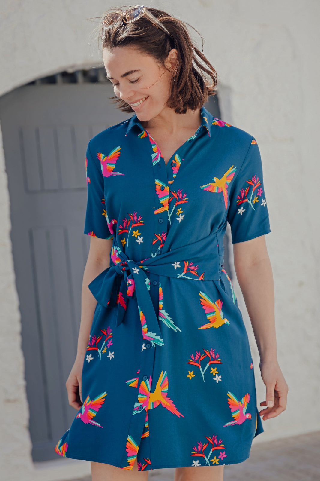 A model wearing a teal shirt dress with a Rainbow Parrot print, featuring a button-up front, short sleeves, and a tie-front belt.
