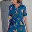 A model wearing a teal shirt dress with a Rainbow Parrot print, featuring a button-up front, short sleeves, and a tie-front belt.